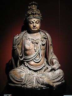 A wooden carving of a slightly overweight Buddha, sitting in a cross legged position. Clothing, including a shirt that covers the shoulders but leaves the chest exposed, and long, baggy pants, are carved into the statue.
