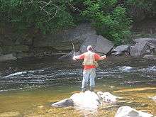 A man in an orange shirt and tan vest fishes in a stream with rapids and large boulders