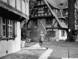 A monochrome photograph of a large man walking away from the viewer, accompanied by a small dog, walking between two buildings with half-timbered architectural styling.