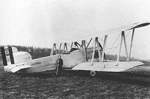 A monochrome photograph of a biplane parked on an airfield, with a man posed leaning against its fuselage with his hands in his pockets