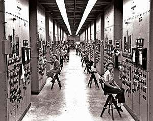 A long corridor with many consoles with dials and switches, attended by women seated on high stools.