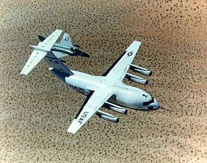 Top view of cargo aircraft in-flight, trailed by a fighter chase aircraft. Under each un-swept wing are two engines suspended forward ahead the leading edge.