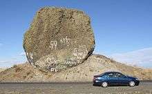 This photo shows an automobile passing in front of a rock which is essentially fully exposed. The rock has a rough, dark surface indicating it is weathered basalt and is roughly circular in exposed cross-section. The rock is immediately adjacent to a roadway – the road cut removed much of the earth from one side of it exposing it – from the excavation it is evident that the rock sits on a mound of glacial till. The rock is approximately 2 times the length of the car (i.e., ~ 9 meters) in one direction and 5 times the height of the car in the other direction (i.e., ~ 9 meters). Since the rock has not tipped onto the road and no structural support is provided, it must be approximately as deep as it is wide and high. Since the density of basalt is 3 grams per cubic centimeter, this puts the mass of the rock at about 400 to 500 metric tons (consistent with the references).