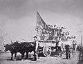Wagon being pulled by oxen, with large number of people standing in the wagon, one holding a large flag