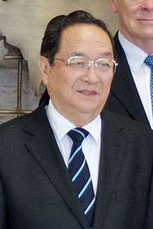 a nearly bald man wearing glasses, a white shirt, a suit and a black tie with blue stripes
