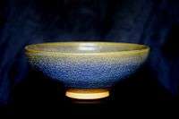 A ceramic bowl is viewed from one side. The glaze has a crazed and mottled finish, varying from yellow at the rim of the bowl to blue below.