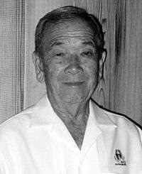 Head and shoulders of an elderly man wearing a white button shirt with an emblem on the left breast