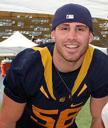 American football player standing with arms akimbo, in gold jersey (#56) and dark blue pants, wearing a dark blue helmet
