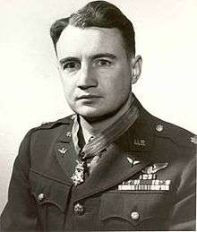 Head and shoulders of a man with wavy hair wearing a military jacket with rows of ribbon bars and a winged pin on his left breast, pins on the lapels, and a star-shaped medal hanging from a ribbon around his neck.