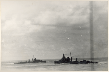 A dark gray warship cruises at high speed while another destroyer steams in the opposite direction