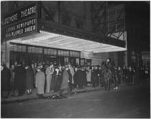 Black and white photo of a crowd of people standing on the sidewalk in front of a building that has a lighted marquee saying 'Biltmore Theatre'.